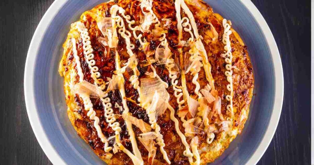 A golden-brown Chicken Okonomiyaki topped with vibrant green onions, savory sauce, and a sprinkle of sesame seeds, served on a traditional Japanese plate. easy chicken okonomiyaki Easy Chicken Okonomiyaki Easy Chicken Okonomiyaki 1024x538
